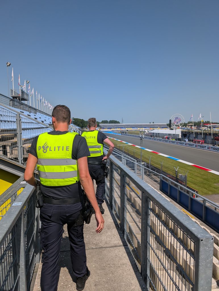 Two police officers in high-visibility vests walk along a metal railing beside a race track, with stands and flags to the left and a ferris wheel in the distance on the right.