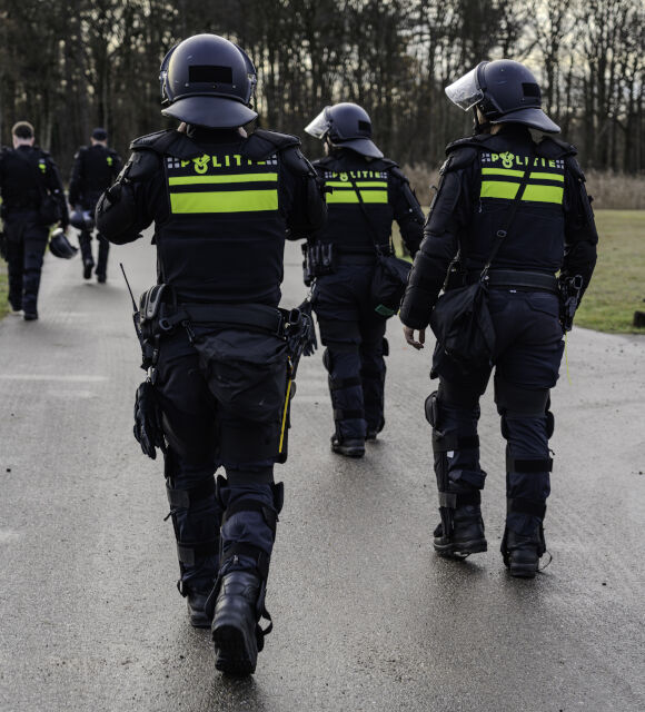 Group of police officers in riot gear walking away on a damp road, backs to the camera, helmets with visors and bright yellow a‑stripe jackets
