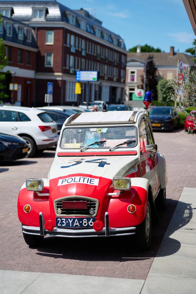 Front of a white and red Dutch police car with a blue emergency light on the roof, license plate 23-YA-86, parked on a brick street with buildings and other cars in the background.