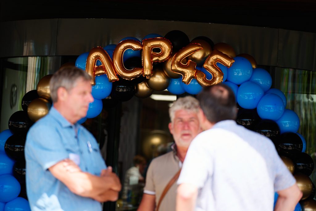 Gold foil balloons spell the word 'ACES' arranged among blue and black balloons at a doorway, with people standing in front.
