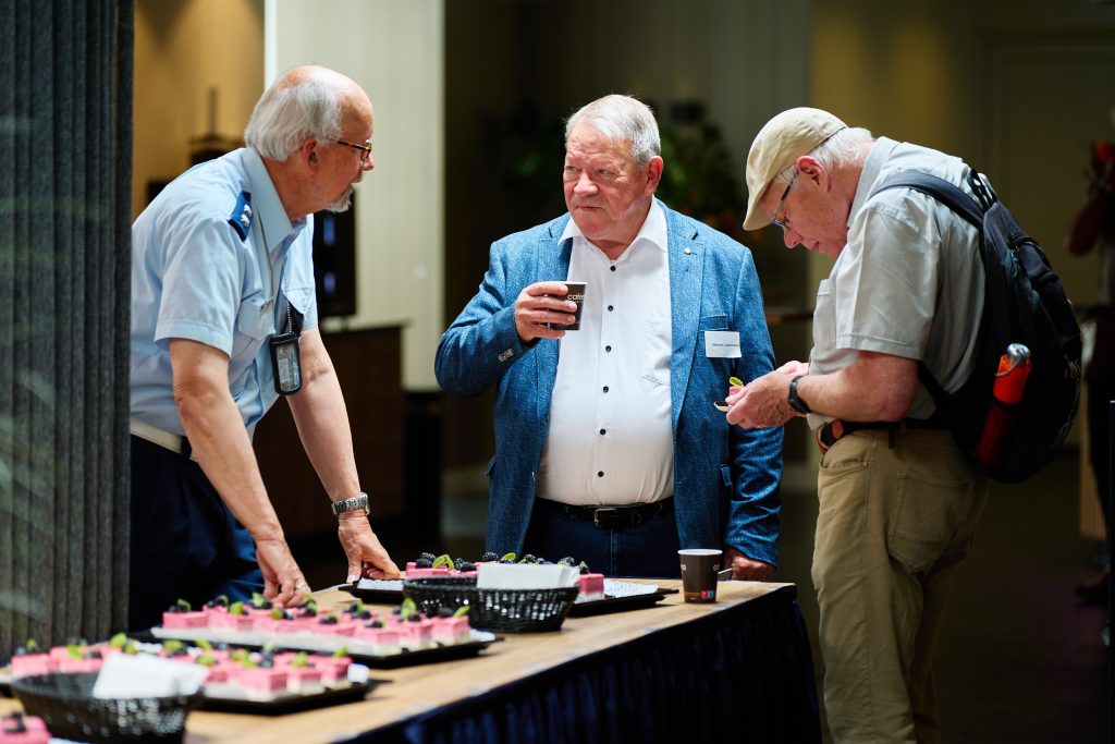 Three older men stand around a table with pink-frosted cupcakes, one holding a cup of coffee.