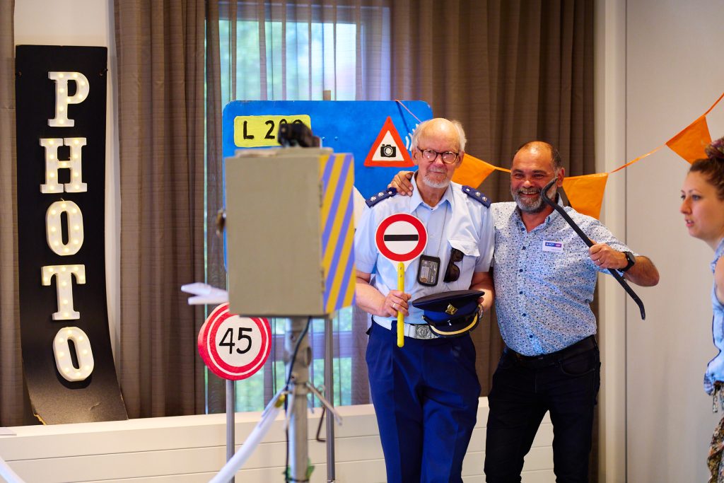 Two men posing with road signs and props at an indoor event, one in a police-style uniform, both smiling, orange bunting in the background.