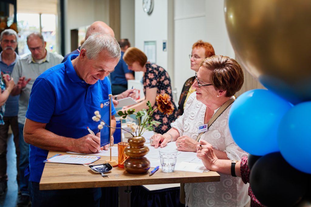 Man in a blue polo signs a document at a registration desk while a woman in a white lace blouse watches, with flowers and balloons in view at a social event.