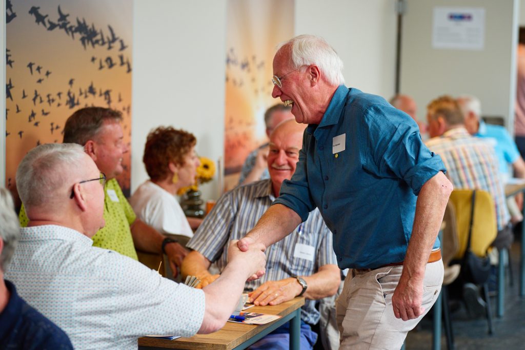 Older man in a blue shirt shakes hands with seated guests at a long table during a social gathering.