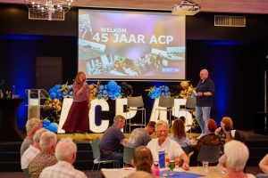 Two hosts on a decorated stage at ACP's 45th anniversary, with a large screen reading 'WELKOM 45 JAAR ACP' and blue‑gold balloons behind them.
