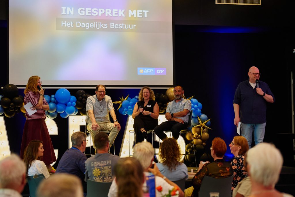 Panel discussion on a stage with five people seated, microphones in hand, audience watching; colorful balloons decorate the background and a projection screen reads Dutch text.