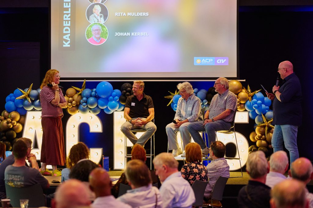 Female host with a microphone standing left on a lit stage, speaking to a panel of five, with blue and gold balloon decorations and large illuminated letters in the foreground; projector screen shows speakers Rita Mulders and Johan Kerbel.