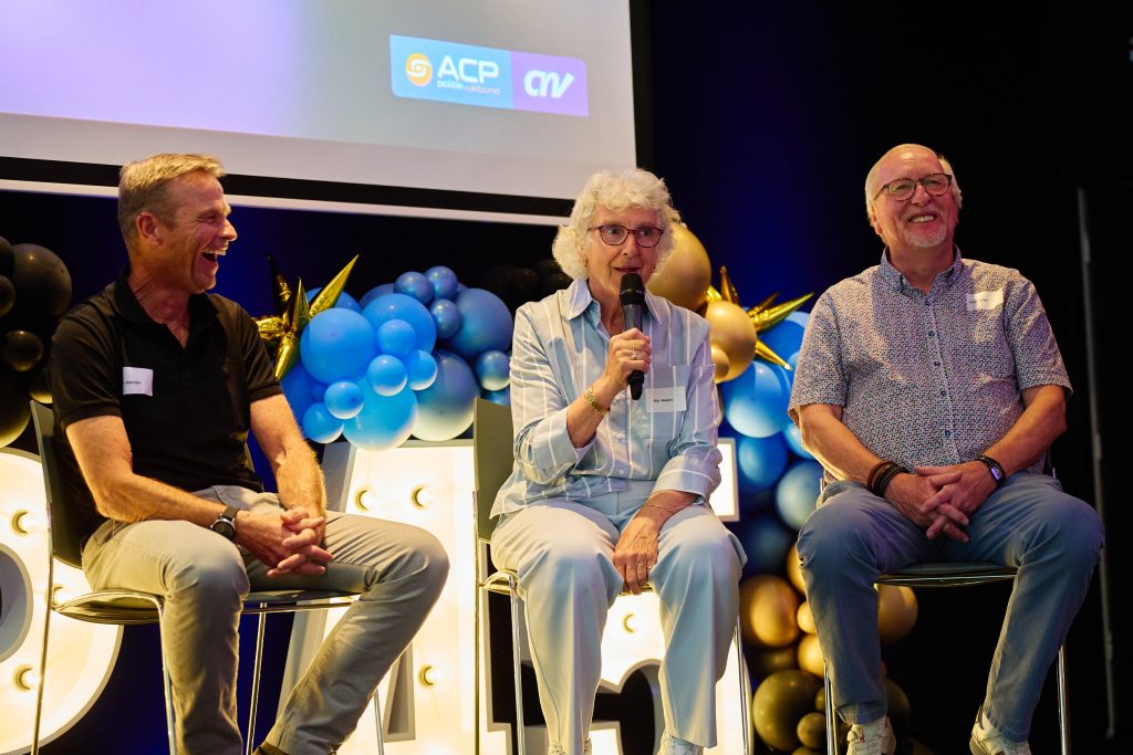 Panel of three people smiling on stage during a discussion, with blue balloon decor and a projection screen showing logos in the background.