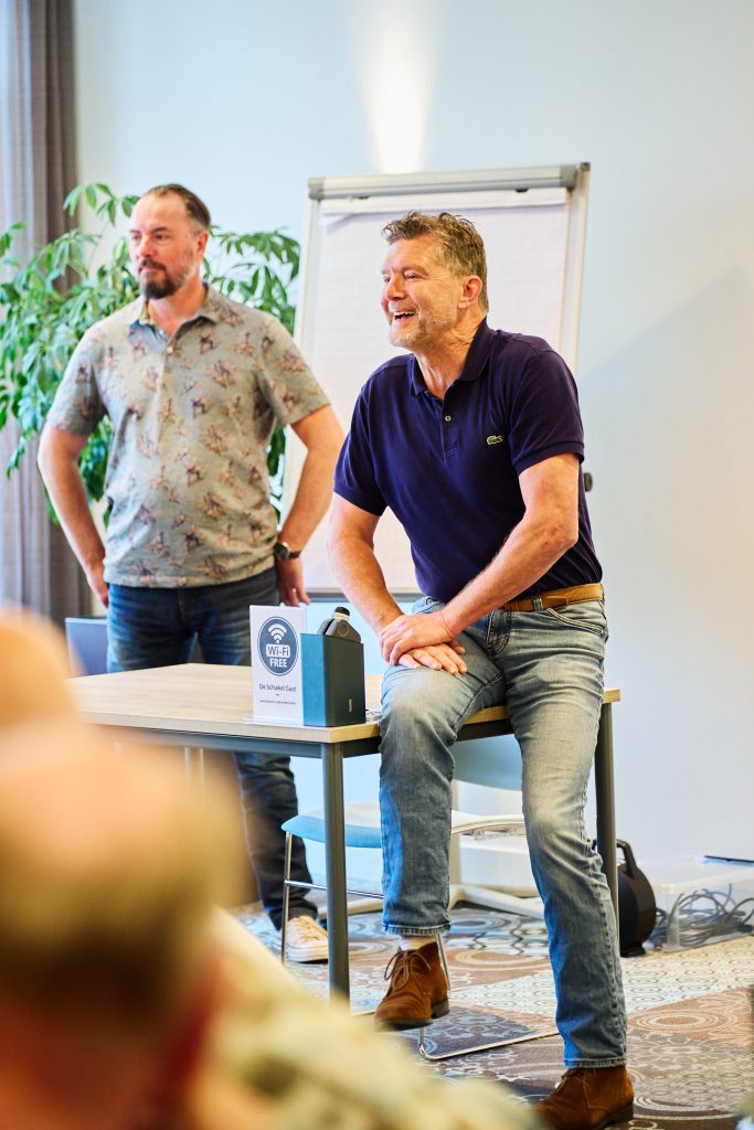 Two men presenting in a bright room; one in a patterned shirt, the other in a navy polo, near a flip chart with a plant in the background.