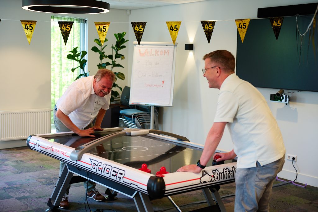 Two men playing air hockey at a table in an office-like room with yellow and black '45' pennant banners overhead, cheering a game moment.