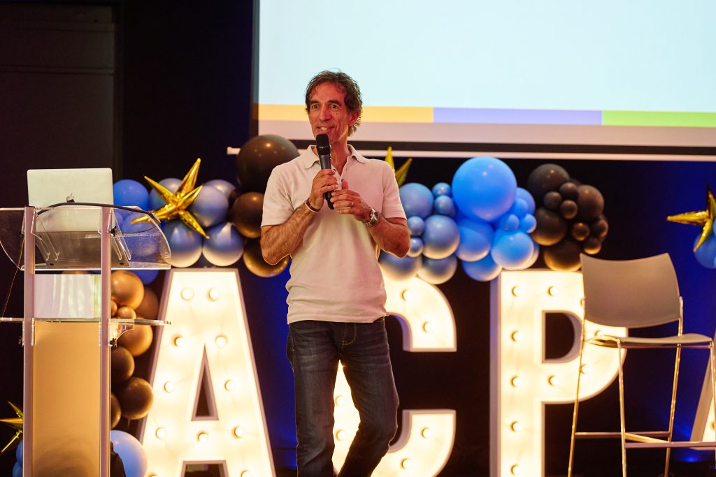 Man in a light polo shirt holding a microphone, speaking on a decorated stage with balloons and illuminated letters behind him.