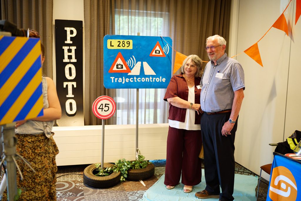 Two smiling older adults posing beside a blue road-safety display labeled 'Trajectcontrole' with triangular traffic signs and orange bunting in the background.