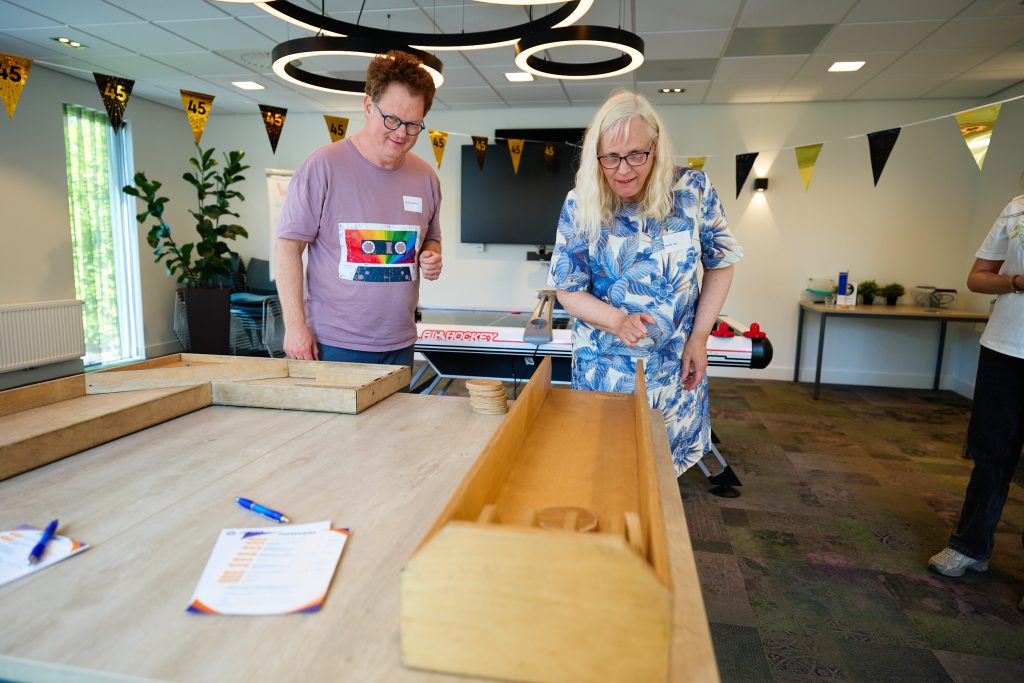Two adults wearing name tags stand at a table with wooden frames, looking down at a game setup in a bright event space with gold and black bunting overhead.