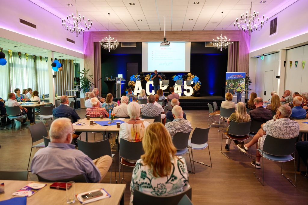 Audience seated at round tables in a decorated conference hall, watching a speaker on stage with a projection screen and balloon decorations nearby.