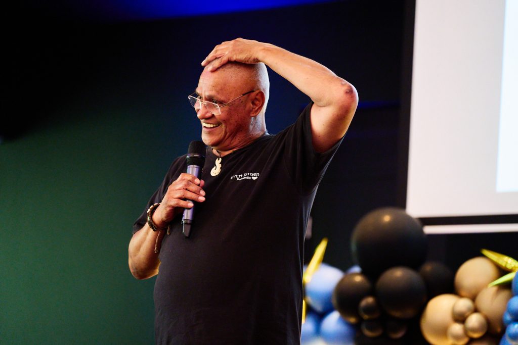 Speaker with a microphone, smiling, hand on head during a talk at an event with balloons in the background.