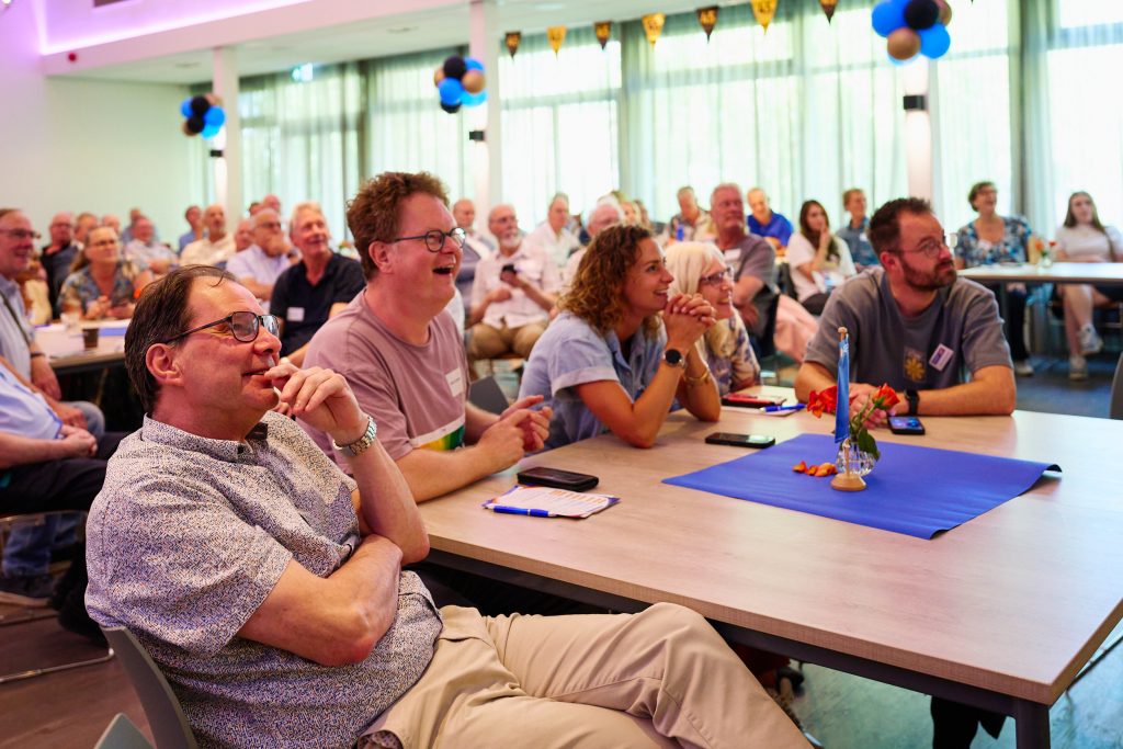 Audience of adults seated at tables watching a presentation in a bright conference room with balloons overhead.