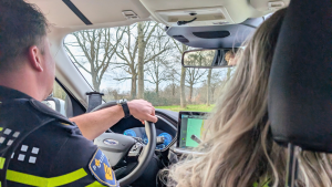 Interior of a police car: officer driving with hand on the steering wheel, GPS/navigation screen on the dash, passenger with long hair in the foreground, trees visible outside the windshield.