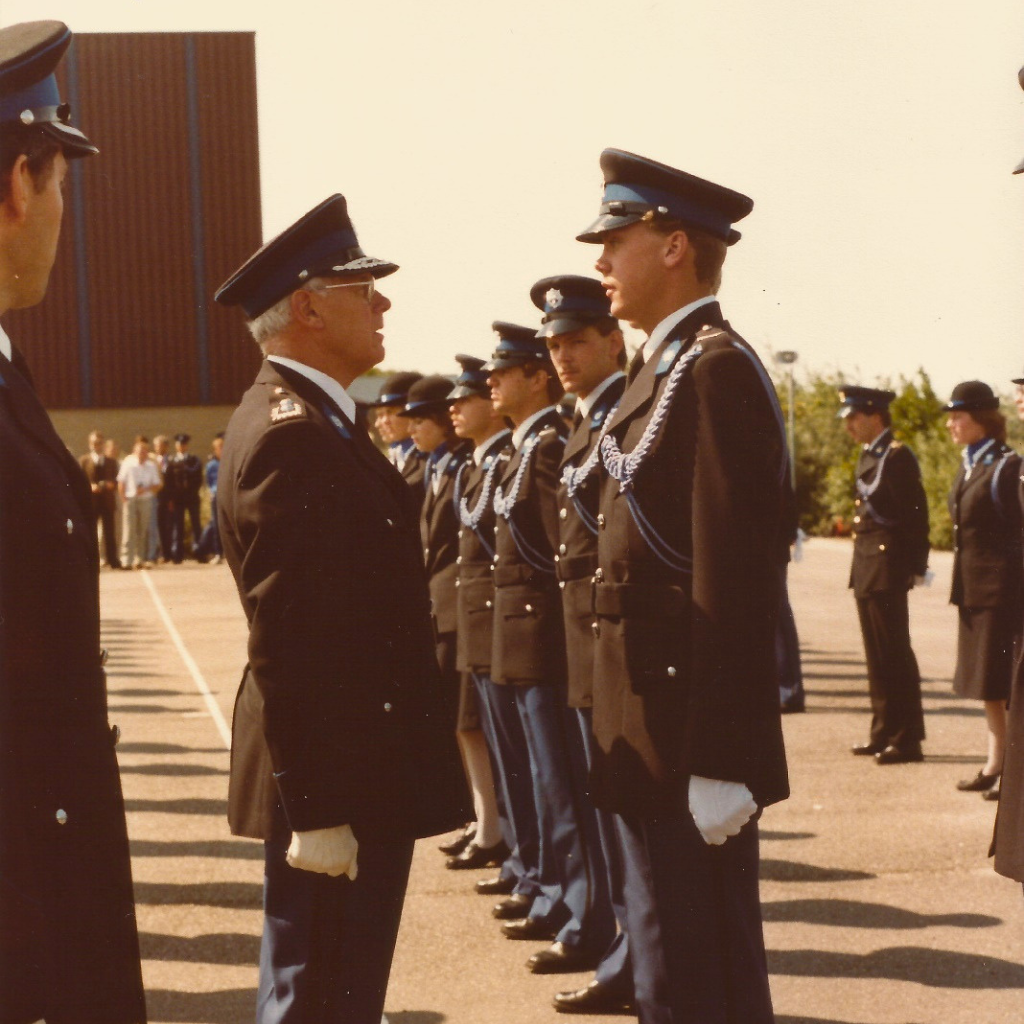 An older officer in a dark dress uniform faces a younger officer in formal uniform, with a line of similarly dressed troops in formation behind them on a sunny day.
