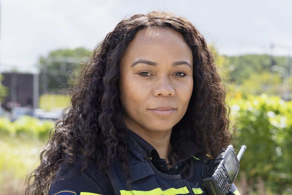 Female firefighter in turnout gear outdoors with a radio on her chest, facing the camera.