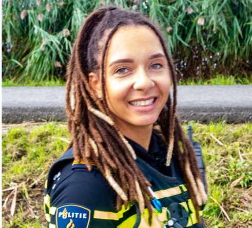 Female police officer in a dark uniform with yellow reflective stripes, smiling outdoors.