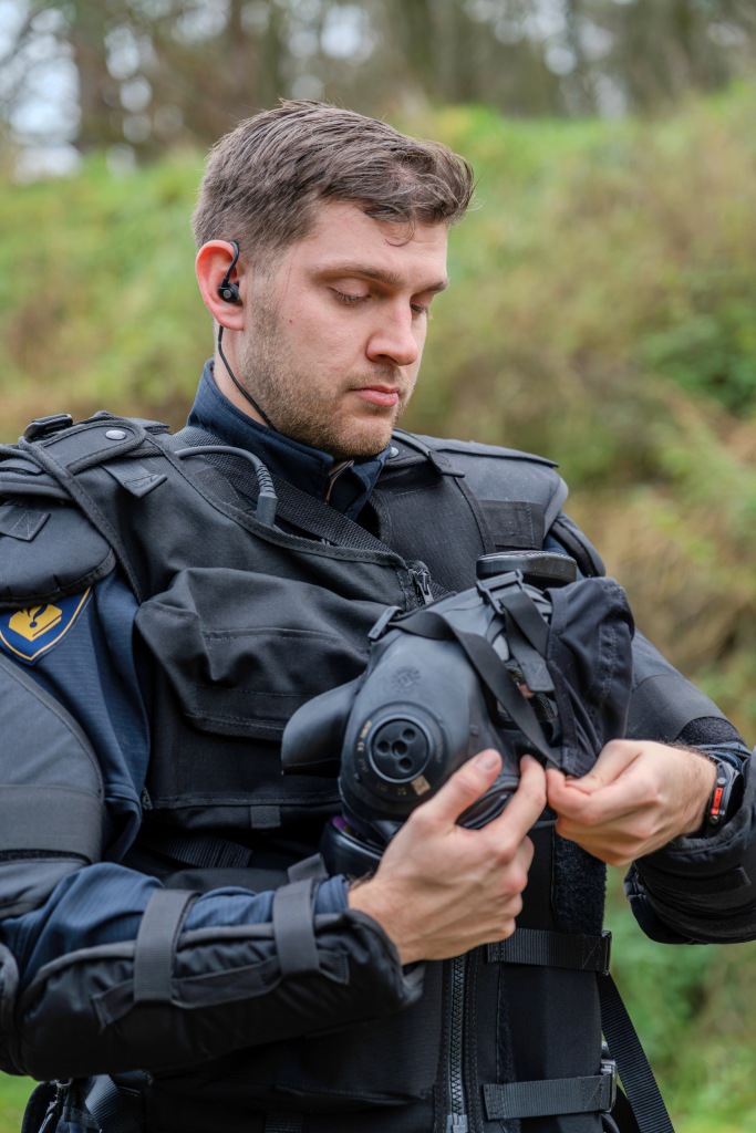 Man in a black tactical vest outdoors, wearing an earpiece and holding a gas mask.