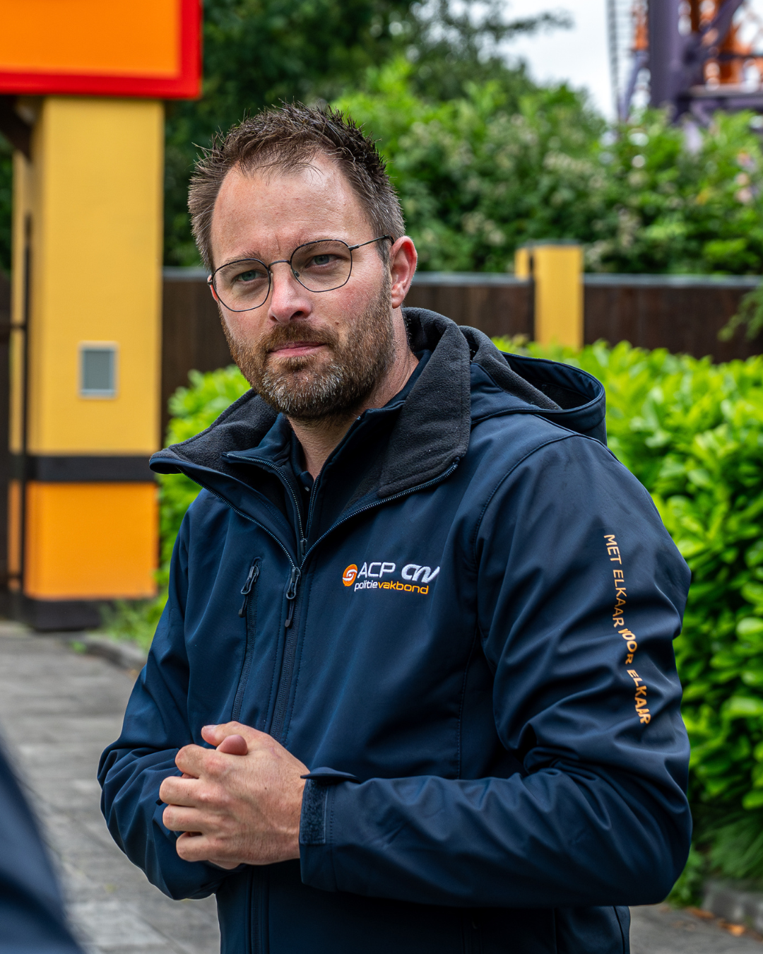 Portrait of a man with glasses and stubble wearing a navy jacket with a logo, standing outdoors with greenery in the background.