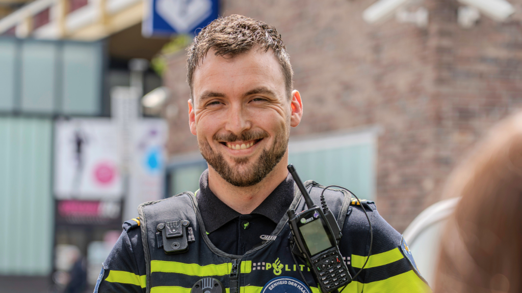 Smiling firefighter in a dark uniform with reflective yellow stripes, a radio clipped to his vest, standing outdoors and looking at the camera.