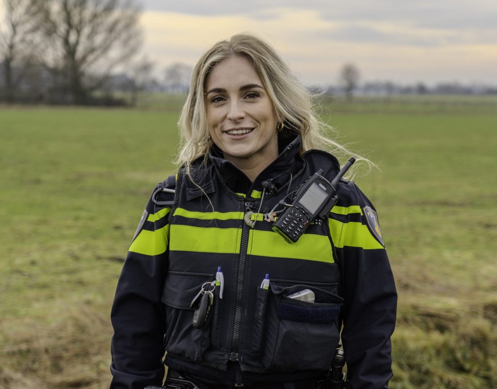 Smiling woman in a black-and-yellow high-visibility jacket with radio and gear, standing in a grassy field outdoors.