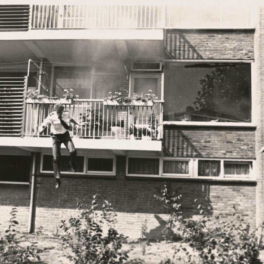 Man jumping from a second-floor balcony of a brick apartment building, arms outstretched in midair.