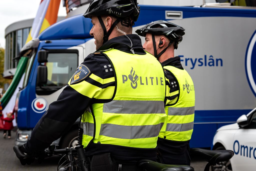 Two Dutch police officers on bicycles in neon yellow jackets, with a blue police van labeled Fryslân in the background next to them.