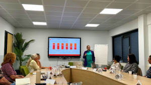 Woman presenter in teal and purple stands by a screen showing a bar chart to a round-table group in a conference room.