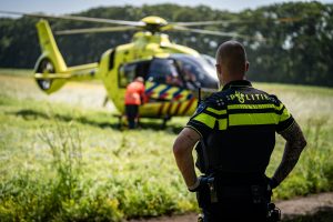 Back view of a police officer in a neon jacket watching a yellow emergency helicopter with its door open on a grassy field behind him.