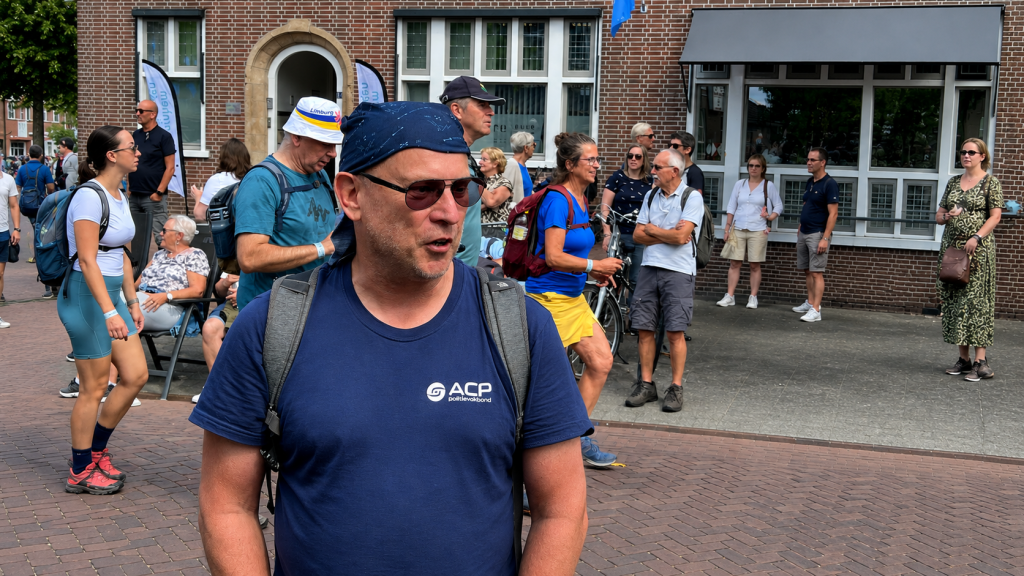 Man with sunglasses and a blue t-shirt with a backpack speaks to others in a crowded outdoor event next to a brick building.