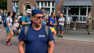 Man with sunglasses and a blue t-shirt with a backpack speaks to others in a crowded outdoor event next to a brick building.