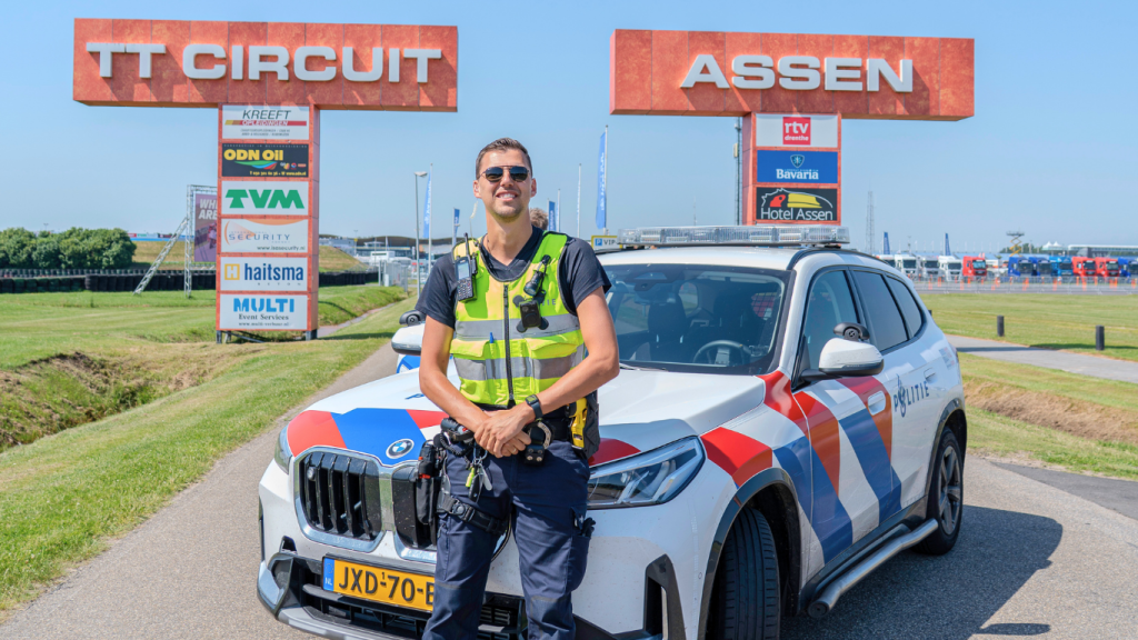 Dutch police SUV with blue-red chevrons and a smiling officer in a high-visibility vest stands in front of the car at TT Circuit Assen, with large red TT Circuit and Assen signs in the background and sponsor boards nearby.