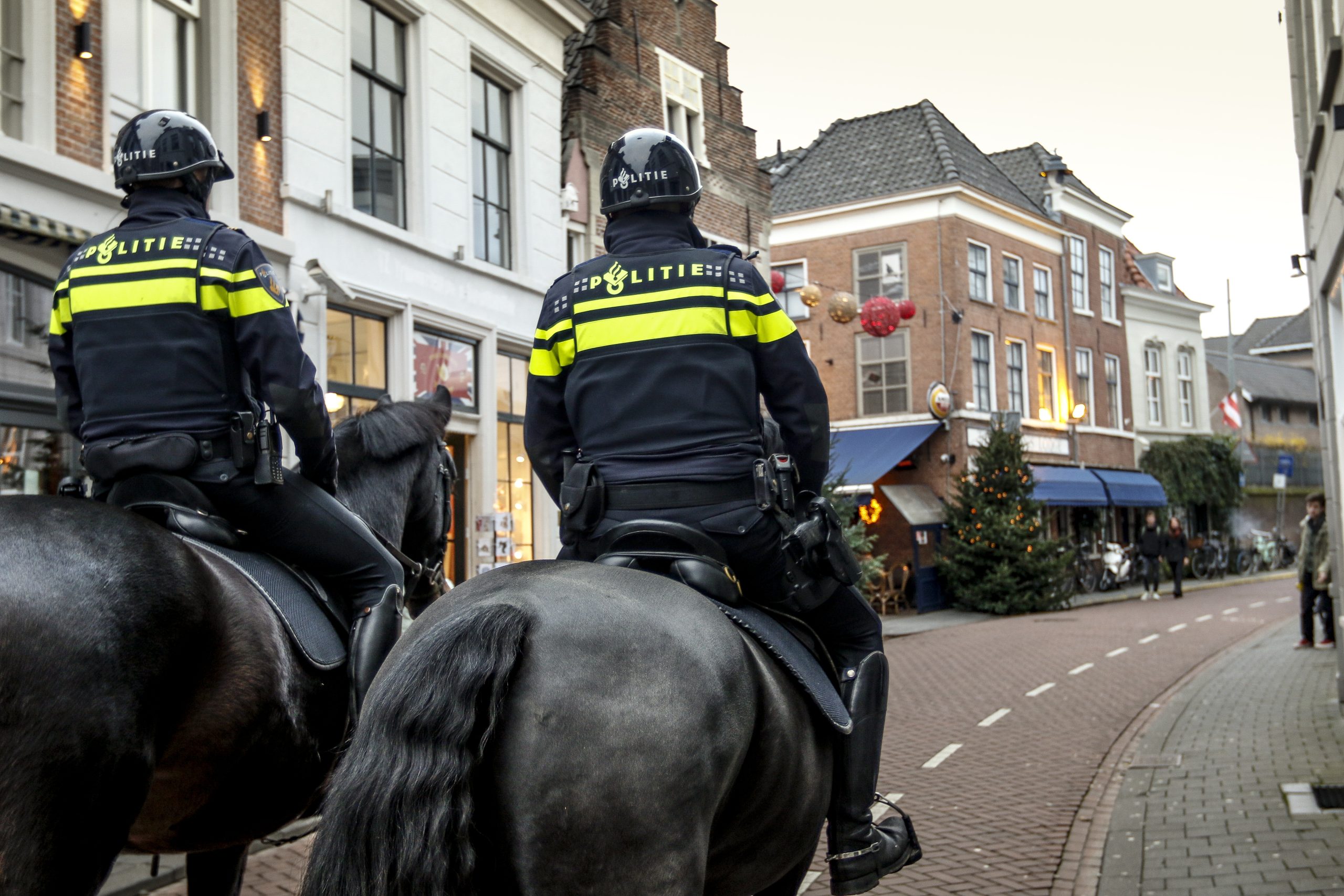 Two police officers on horseback patrol a pedestrian street in a European town, seen from behind.